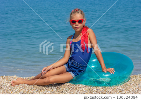 A girl in a one-piece swimsuit with red braids and red sunglasses, sitting gracefully on a green inflatable ring on a pebble beach. 130324084