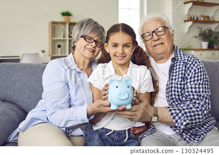 Girl granddaughter sitting with her senior grandparents on sofa at home with piggy bank. Girl granddaughter sitting with her senior grandparents on sofa at home with piggy bank. 130324495