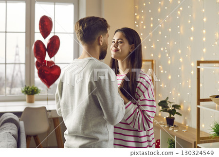 Man and woman stand and look at each other with loving eyes in romantic setting in living room at home. Happy, beautiful, young couple celebrate Valentine's day, birthday, holiday. Man and woman stand and look at each other with loving eyes in romantic setting in living room at home. Happy, beautiful, young couple celebrate Valentine's day, birthday, holiday. 130324549