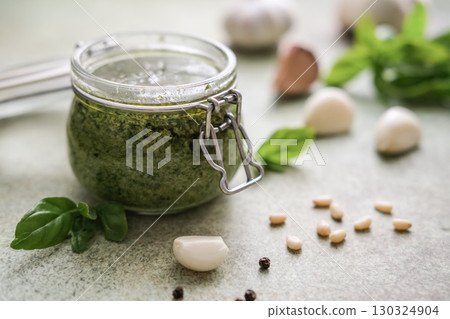 Fresh homemade basil pesto in a glass jar, surrounded by ingredients like garlic, pine nuts and fresh basil leaves on speckled background 130324904