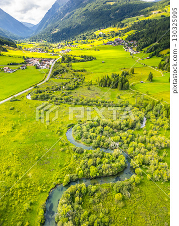 A winding river flows through a lush green valley surrounded by mountains. The vibrant landscape features farms, trees, and a quaint village in the distance. 130325995