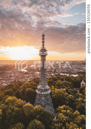 Petrin Lookout Tower stands majestically as the sun rises, casting a warm glow over the surrounding hills and vibrant tree canopy in Prague, revealing the city's beauty at dawn. 130326056