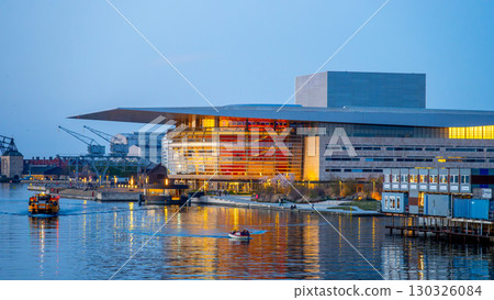 Copenhagen Opera House illuminates the evening sky with vibrant colors, reflecting beautifully on the water. Boats glide by as the cityscape provides a stunning backdrop in Denmark. Copenhagen Opera House illuminates the evening sky with vibrant colors, reflecting beautifully on the water. Boats glide by as the cityscape provides a stunning backdrop in Denmark. 130326084