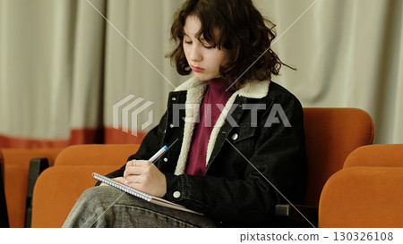 A focused girl sits in an auditorium chair, immersed in her notebook writing. The scene captures the dedication and introspection involved in personal growth and creativity. 130326108