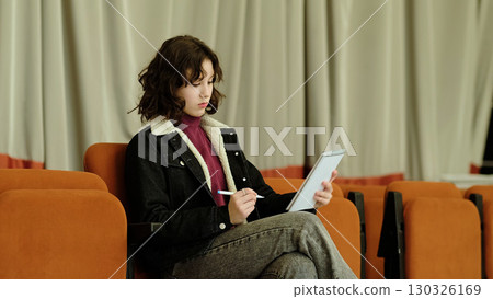 A thoughtful girl writes in a notebook while seated in a quiet auditorium. This image symbolizes themes of education, mindfulness, and the joy of creativity. 130326169