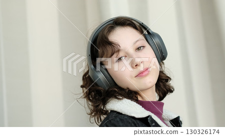 A thoughtful girl with headphones relaxes in a light-filled space, her expression serene. The image emphasizes the personal enjoyment of music and introspective moments. 130326174