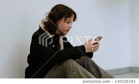 A child in casual attire uses her smartphone while sitting against a blank wall. The photo highlights the pervasive role of technology in everyday life and youth s engagement with digital devices. A child in casual attire uses her smartphone while sitting against a blank wall. The photo highlights the pervasive role of technology in everyday life and youth s engagement with digital devices. 130326191