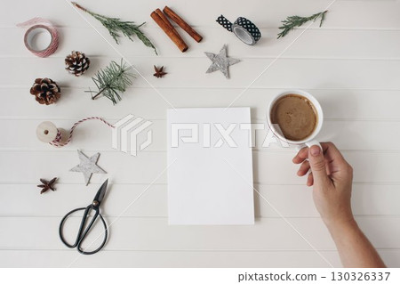Christmas decorations flatlay. Pine cones, cinnamon, fir branches, ribbon scissors. White wooden table background. Blank card, invitation mockup. Hand holding coffee. Festive banner. Holiday greetings 130326337