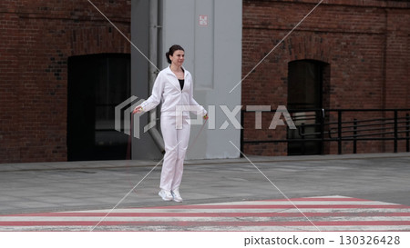 A woman in a white tracksuit jumps rope on an urban crosswalk, focused on her workout. The mix of fitness and city architecture reflects the growing trend of integrating exercise into daily routines. 130326428
