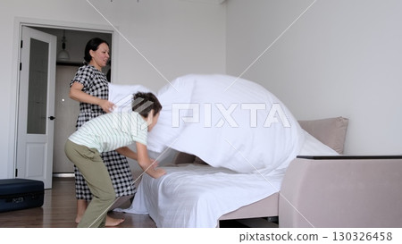 A mother lifts a bedsheet in the air while her child joyfully jumps on the bed. This playful moment captures the warmth and joy of family life, emphasizing the importance of bonding and shared fun at A mother lifts a bedsheet in the air while her child joyfully jumps on the bed. This playful moment captures the warmth and joy of family life, emphasizing the importance of bonding and shared fun at 130326458