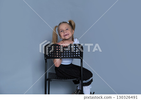 fun moment from a children's fashion shoot, where a 5-year-old girl, dressed in a trendy outfit, sits playfully on a chair. Her bright laughter and energetic pose make this image perfect for kids 130326491