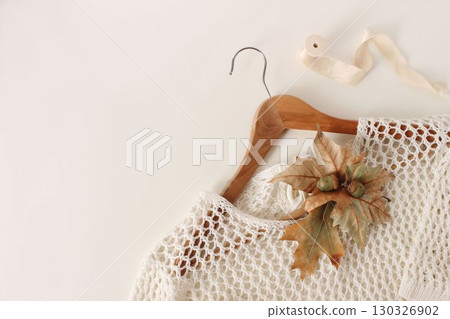 White knitted sweater on wooden hanger decorated with maple plane leaves, acorns. Minimal cozy autumn composition, natural elements. Fall seasonal fashion, lifestyle. Flatlay, top view, copy space 130326902