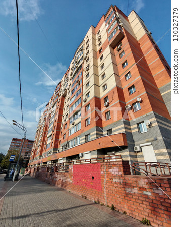 A tall red and beige brick residential building with multiple balconies, air conditioning units, and a sidewalk below. The structure reflects the typical architecture of urban apartment blocks in 130327379