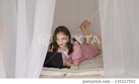 A young woman works on a laptop in a cozy bedroom surrounded by soft white curtains. This image reflects the growing trend of remote work and balancing productivity with comfort. 130327501