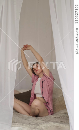 A woman stretches gracefully on a comfortable bed, basking in the calm of her surroundings. The photo reflects the beauty of slow mornings and prioritizing well-being in daily life. 130327502