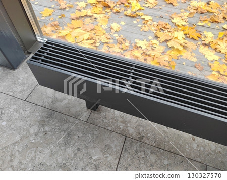 A modern floor heating vent near a glass window with autumn leaves scattered outside. The photo combines indoor functionality with the charm of seasonal outdoor scenery. 130327570