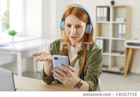 Shocked young woman in wireless headphones holding phone, cellphone user girl staring at screen Shocked young woman in wireless headphones holding phone, cellphone user girl staring at screen 130327719