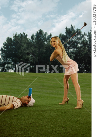 Woman golfer in pink dress swinging club near man lying with soda can on head bold playful mood 130327789