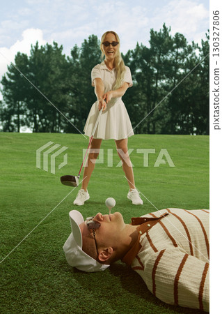 Woman golfer laughing while aiming golf club at man lying with ball on mouth playful risky mood 130327806