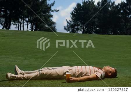 Male golfer lying on grass holding beer glass with relaxed expression peaceful vibe 130327817