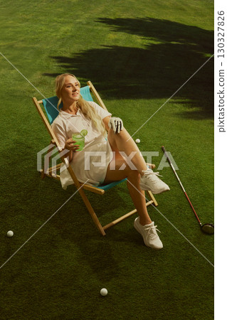 Female golfer relaxing in chair on green grass holding cocktail with joyful smile Female golfer relaxing in chair on green grass holding cocktail with joyful smile 130327826