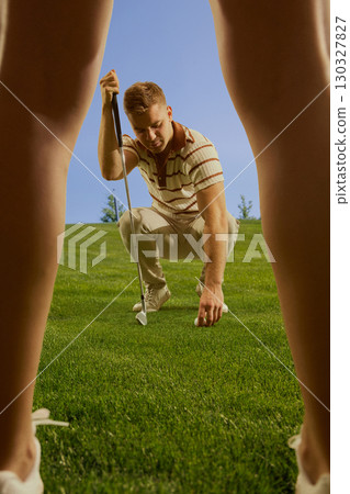 Male golfer crouching to place ball on grass seen through female legs focused confident mood Male golfer crouching to place ball on grass seen through female legs focused confident mood 130327827