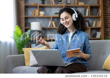Happy female freelancer wearing headphones and holding notebook is speaking at online meeting using laptop while sitting on sofa in cozy living room. Happy female freelancer wearing headphones and holding notebook is speaking at online meeting using laptop while sitting on sofa in cozy living room. 130328118