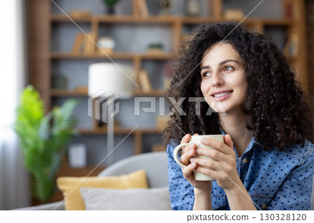 Young woman with curly hair holding a mug and relaxing on the sofa, enjoying a moment of peace and tranquility in her modern living room, smiling and looking away. 130328120