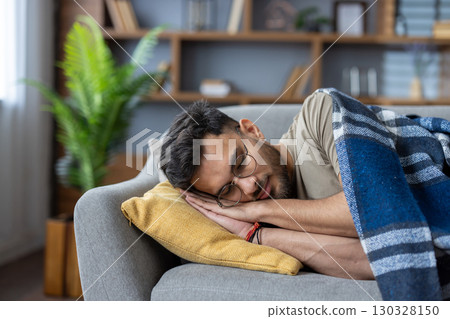 Indian young man wearing glasses is sleeping on the sofa at home, resting on a pillow and covered with a blue blanket. Close-up photo. 130328150