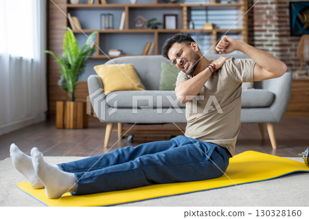 Man sitting on yoga mat stretching arm, clearly feeling shoulder pain in comfortable living room. Indoor exercise scene, emphasis on discomfort and injury during physical activity. Man sitting on yoga mat stretching arm, clearly feeling shoulder pain in comfortable living room. Indoor exercise scene, emphasis on discomfort and injury during physical activity. 130328160