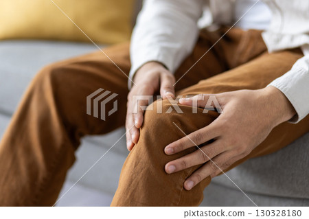 Person seated on sofa holding knee, indicating discomfort or pain. Captures concept of injury, health concern, or joint problem. Emphasizes human touch and care related to knee pain. Person seated on sofa holding knee, indicating discomfort or pain. Captures concept of injury, health concern, or joint problem. Emphasizes human touch and care related to knee pain. 130328180