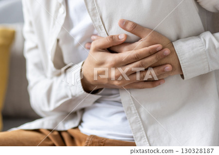 Close-up of person holding stomach with hands indicating discomfort or pain. Individual is dressed in casual attire, seated indoors, suggesting potential issues like indigestion, cramps 130328187