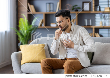 Muslim young man sitting on the sofa at home, holding his chest with his hand, feeling severe pain, coughing and covering his mouth with his hand. Muslim young man sitting on the sofa at home, holding his chest with his hand, feeling severe pain, coughing and covering his mouth with his hand. 130328193