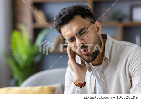 Young man wearing glasses holding his ear with pained expression. He appears to be experiencing ear discomfort, possibly due to an earache. Indoor home background suggests casual environment. Young man wearing glasses holding his ear with pained expression. He appears to be experiencing ear discomfort, possibly due to an earache. Indoor home background suggests casual environment. 130328199