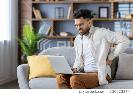 Contorted in pain, a young Indian man is at home, working on a laptop, sitting on a sofa and holding his hand behind his back. 130328274
