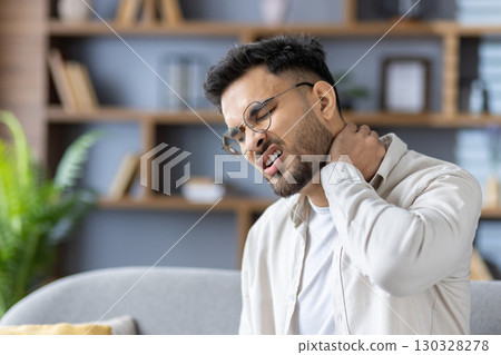 Man holding neck with pained expression, indicating discomfort and stress. Sitting on couch in living room setting, conveying concept of physical pain or injury, possibly due to posture or stress. Man holding neck with pained expression, indicating discomfort and stress. Sitting on couch in living room setting, conveying concept of physical pain or injury, possibly due to posture or stress. 130328278