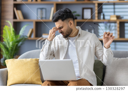 Man sitting on couch with laptop experiencing discomfort and eye strain, holding glasses and massaging forehead. Concept of remote work challenges, stress, and digital eye strain. 130328297