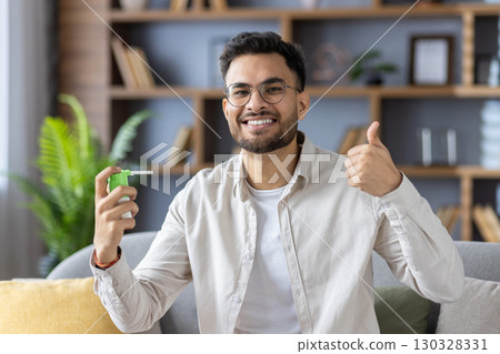 Smiling man with glasses holding throat spray shows thumbs up indicating relief and satisfaction. Person in cozy home setting with modern decor conveys sense of comfort and wellness. 130328331