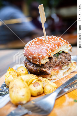 Juicy hamburger with sesame seed bun, garnished with fresh vegetables, served alongside crispy roasted potatoes on wooden platter Juicy hamburger with sesame seed bun, garnished with fresh vegetables, served alongside crispy roasted potatoes on wooden platter 130328360