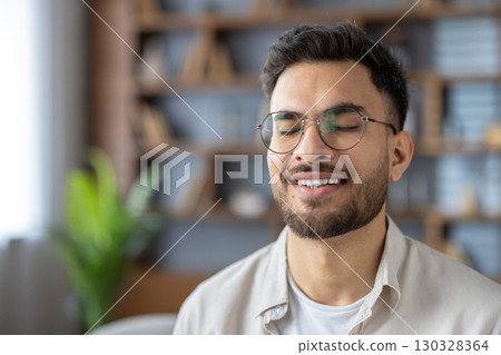 Young man with glasses smiling peacefully while meditating indoors. Capturing moment of relaxation, serenity, and mindfulness. Closed eyes convey sense of calm and inner peace tranquil environment. Young man with glasses smiling peacefully while meditating indoors. Capturing moment of relaxation, serenity, and mindfulness. Closed eyes convey sense of calm and inner peace tranquil environment. 130328364