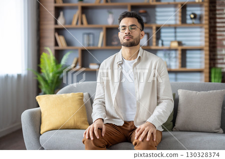Young man sitting on sofa with eyes closed, practicing meditation and mindfulness. He is focusing on relaxation, inner peace, and mental clarity in cozy home setting. 130328374