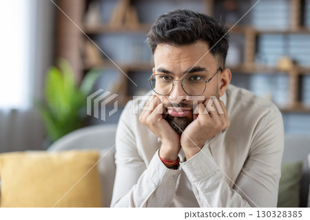 Young man wearing glasses sitting in living room appearing bored and thoughtful. Casual attire highlights relaxed atmosphere. Expression conveys mix of boredom and contemplation 130328385