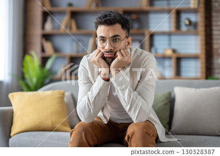 Young man with glasses sitting on sofa, deep in thought, reflecting on life choices. Dressed in casual clothes, conveying introspection and contemplation in relaxing home setting. 130328387