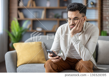 Bored and upset young Indian man sitting on the sofa at home, resting his head on his hand and monotonously looking at his mobile phone. Bored and upset young Indian man sitting on the sofa at home, resting his head on his hand and monotonously looking at his mobile phone. 130328395