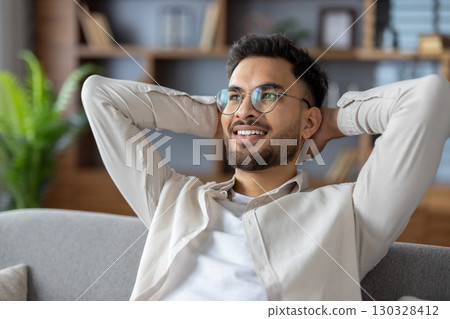 Hispanic man enjoying peaceful moment at home sitting on couch. Smiling with hands behind head, feeling relaxed and content. Capturing happiness and comfort in everyday life. 130328412