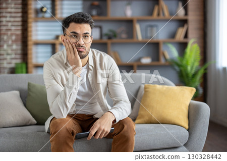 Young man sitting on comfortable sofa holding remote control, appearing bored and disengaged with television content. Casual clothing, glasses, and home setting convey relaxation and lack interest. 130328442
