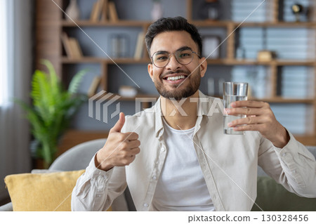 Smiling man with glasses holding glass of water showing thumbs-up. He sits comfortably in modern living room conveying positivity, health, and hydration. Smiling man with glasses holding glass of water showing thumbs-up. He sits comfortably in modern living room conveying positivity, health, and hydration. 130328456