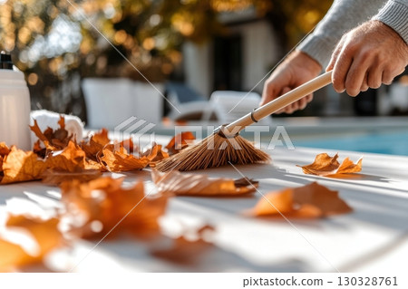 A person sweeps autumn leaves from a patio table near a swimming pool, enjoying a sunny autumn day 130328761