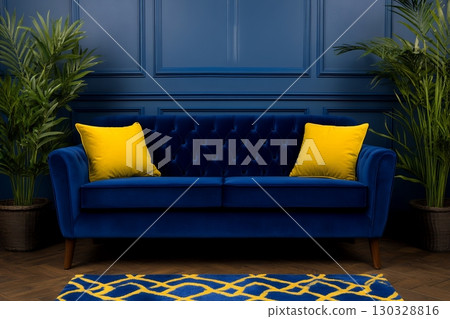 A luxurious blue velvet sofa with yellow cushions sits against a dark blue paneled wall, flanked by potted plants 130328816