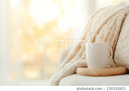 A white mug sits on a wooden tray atop a cozy knitted throw blanket near a bright window 130328831